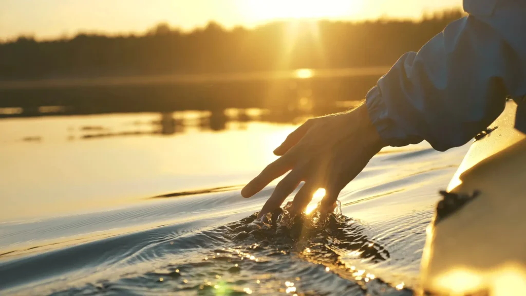Man dips his fingers down in lake Norman while kayaking against backdrop of golden sunset, and closeup of his hand and pristine water.